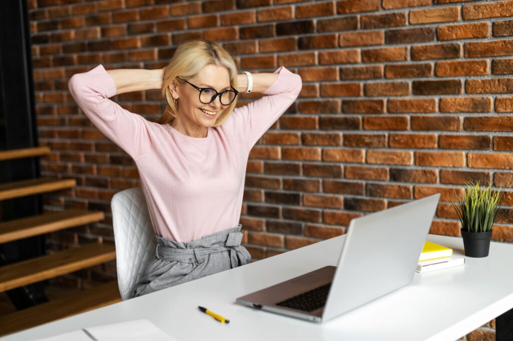 Woman in pink shirt at computer desk stretching with arms behind head